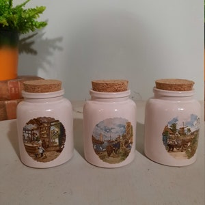 Op de afbeelding: Drie witte keramische potten met kurken deksels en Nederlandse scènes geschilderd op de voorkant. De scènes tonen een winkelinterieur, een haven met boten en mensen, en een windmolen met een paard en wagen.