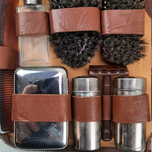 Puede incluir: Un estuche de viaje de cuero marrón con cierre de cremallera. El estuche contiene una variedad de artículos de aseo, incluyendo dos cepillos, un peine, una navaja de afeitar, una botella de aftershave y dos recipientes de metal.