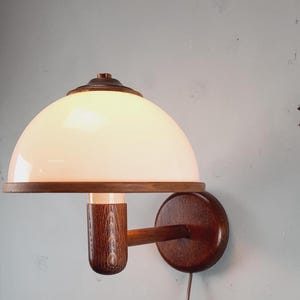 May include: A vintage wall sconce with a white, dome-shaped glass shade and a dark wood frame. The sconce features a round wooden base and a cylindrical wooden support. The light fixture is designed for wall mounting and has a power cord.