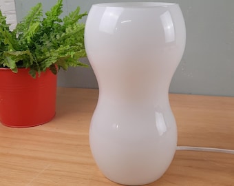 May include: A white, hourglass-shaped glass table lamp. The lamp is smooth and glossy, with a rounded top and base, and a narrower center. A red pot with green plants is to the left.