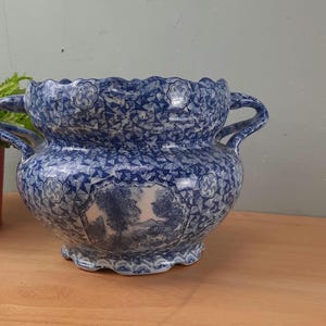 May include: A blue and white ceramic planter with a detailed landscape scene. The planter has two handles and a scalloped rim. The exterior features a repeating geometric pattern. The planter is approximately 25 cm in diameter.