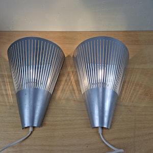 May include: A pair of silver wall sconces with a cone shape. Each sconce features vertical slits, allowing light to pass through. They are connected to a white cable and are mounted on a wooden surface.