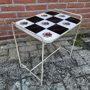 May include: A white metal side table with a black and white checkered top featuring floral designs. The table has a simple, minimalist design.