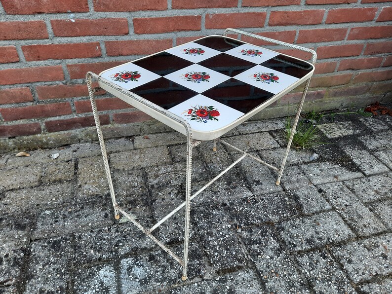 May include: A white metal side table with a black and white checkered top featuring floral designs. The table has a simple, minimalist design.