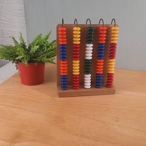 May include: A wooden abacus with colorful beads in red, orange, blue, yellow, green, and white. The abacus is on a wooden surface, with a small red pot of greenery to the left. The abacus is approximately 8 inches tall.