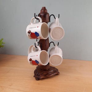 May include: A wooden mug tree holds six white ceramic mugs. The mugs feature a colorful illustration and text. The tree is dark brown with black metal hooks. The mugs are displayed on a wooden table.