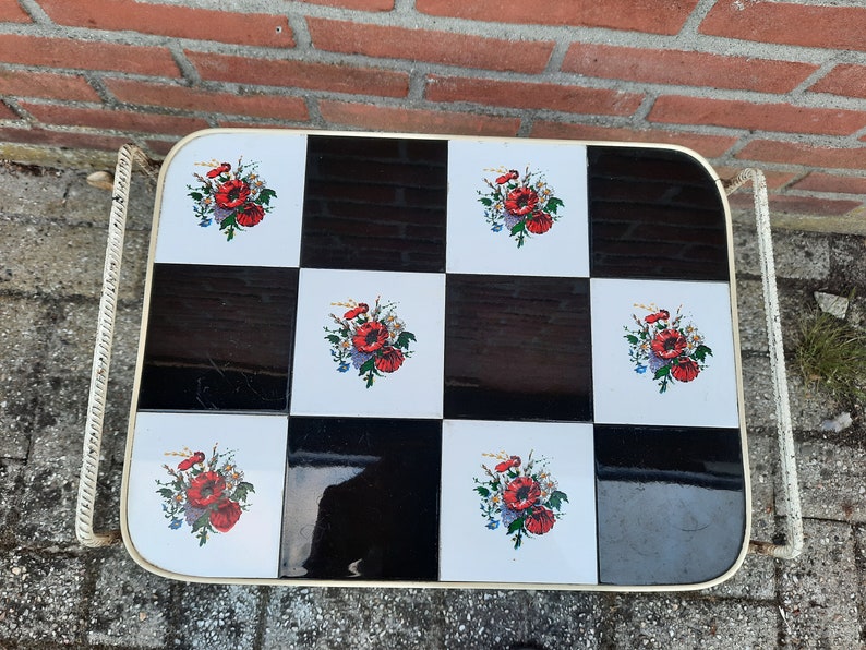 May include: A white metal serving cart with a black and white checkered top. The top is decorated with red and white floral designs.