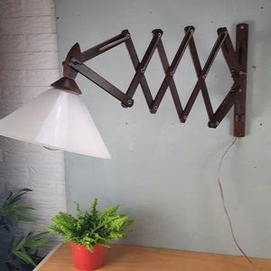 Könnte beinhalten: Eine braune, an der Wand montierte Lampe aus Holz mit einem weißen, kegelförmigen Schirm. Die Lampe hat einen Klapparm und ist an ein Kabel angeschlossen.