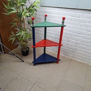 May include: A three-tiered triangular corner shelf unit featuring a green top shelf, a red middle shelf, and a blue bottom shelf. The structure is supported by three vertical legs in matching colors, each topped with a silver-toned cylindrical accent.