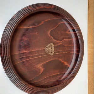 May include: A round wooden serving tray with a dark brown stain. The tray has a raised rim with a decorative pattern and a gold emblem in the centre.