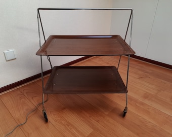 May include: A two-tiered folding serving cart with a chrome frame and brown wooden shelves. The cart has wheels and is on a wooden floor.