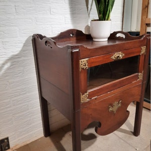 May include: A dark wood side table with a glass door and brass accents. The table has a shelf and a drawer. The table is sitting on a tiled floor.
