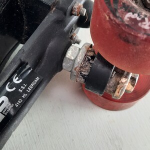May include: Close-up of a black roller skate with a red wheel that says "Othello" on it. The skate has a black metal frame and a silver axle. The skate also has a sticker that says "CE gepr&uuml;ft sicherheit" and "S.S.I. 4143 HL LEERDAM".