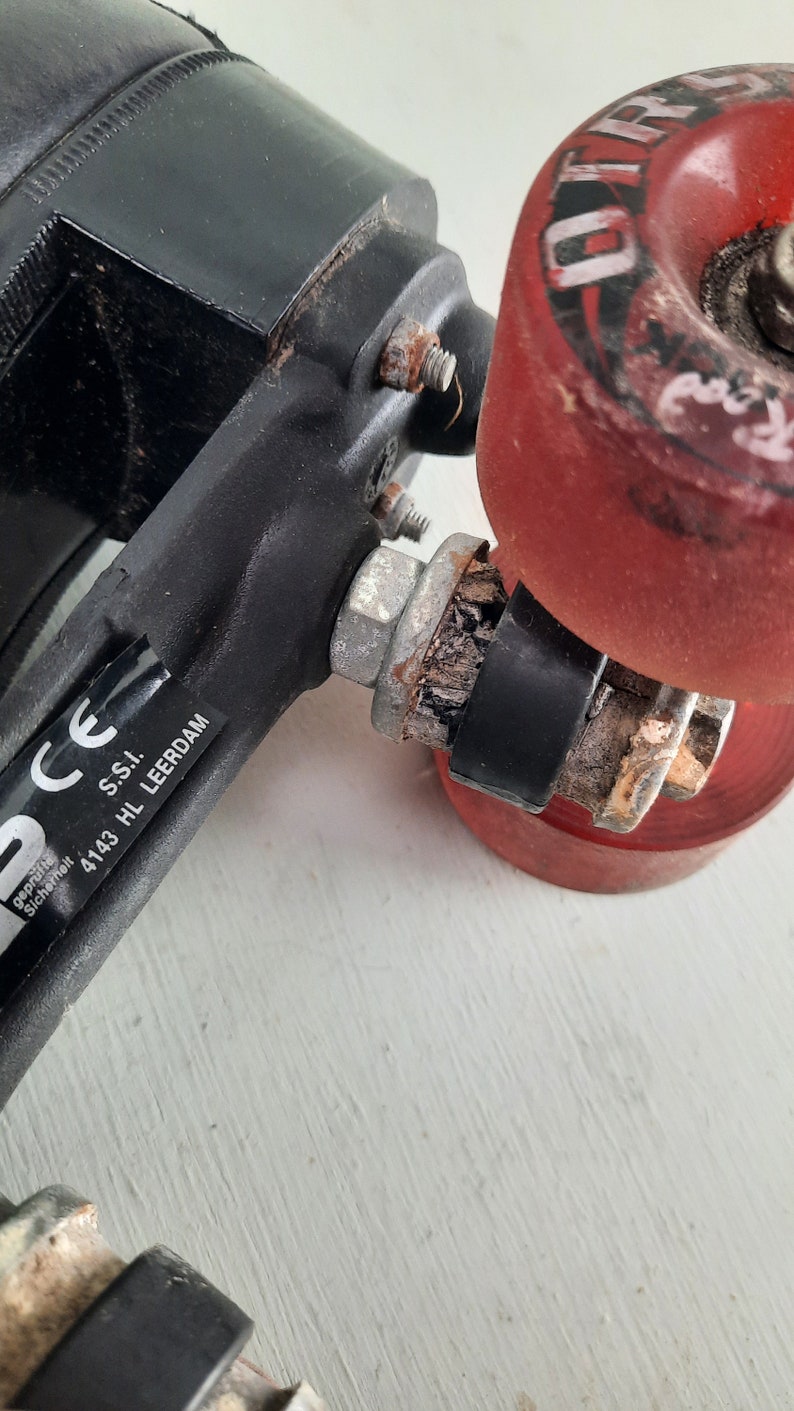 May include: Close-up of a black roller skate with a red wheel that says "Othello" on it. The skate has a black metal frame and a silver axle. The skate also has a sticker that says "CE gepr&uuml;ft sicherheit" and "S.S.I. 4143 HL LEERDAM".