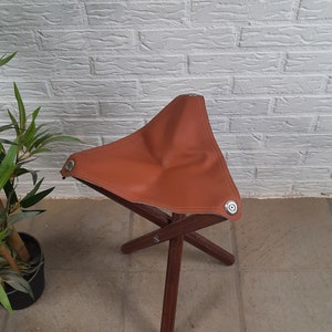 Op de afbeelding: Een bruine leren vouwbare kruk met een houten frame. De kruk is gemaakt van stevig materiaal en heeft een eenvoudig ontwerp.