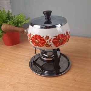 Op de afbeelding: Een vintage fonduepan met een gespikkeld witte buitenkant, versierd met grote oranje bloemmotieven. Het heeft een zilveren deksel en basis, een zwarte standaard en een houten handvat. De pan is ontworpen voor koken op tafel.