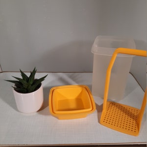 May include: A white ceramic pot with a green succulent plant, two yellow square containers, a clear plastic container with a lid, and a yellow slotted scoop are arranged on a white surface.