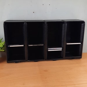 May include: Four black CD storage cases with slots for CDs. The cases are lined up in a row on a wooden surface. Three of the cases have CDs inside.