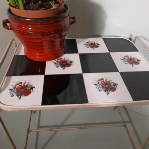 May include: White metal side table with a black and white checkered top. The top is decorated with floral designs featuring red poppies. A red terracotta pot with a plant is sitting on the table.