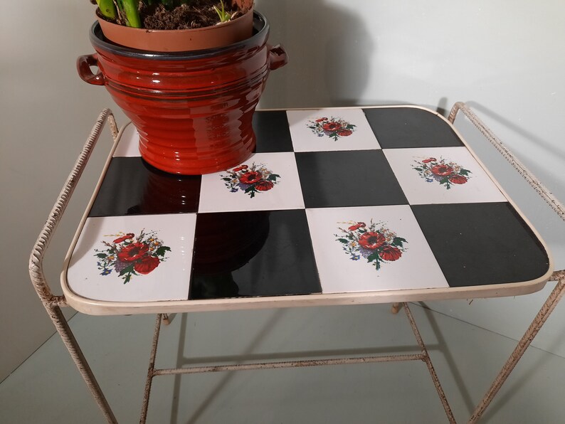 May include: White metal side table with a black and white checkered top. The top is decorated with floral designs featuring red poppies. A red terracotta pot with a plant is sitting on the table.