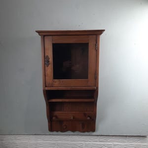 May include: A small, wooden wall cabinet with a glass-paned door, two shelves, and three pegs. The cabinet is made of stained wood and has a decorative metal latch. The cabinet is mounted on a light gray wall.