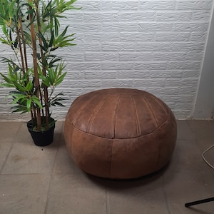 May include: A brown leather pouffe with stitched detailing sits on a tiled floor in front of a white brick wall.