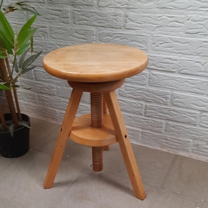 Op de afbeelding: Een houten verstelbare kruk met een ronde zitting en drie poten. De kruk is gemaakt van lichtbruin hout en heeft een schroefmechanisme om de hoogte aan te passen.