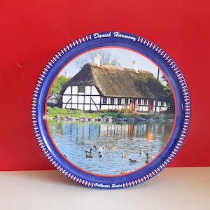 Op de afbeelding: Een ronde blauwe en rode blik met een wit en zwart huis met rieten dak en een vijver met eenden ervoor. De blik heeft de tekst "Danish Harmony Collector Series" op de bodem.