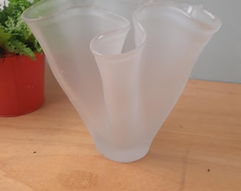May include: A frosted glass vase with a unique, wavy design. The vase has a wide opening and a flared, sculptural shape. The frosted glass gives it a soft, diffused appearance. The vase is sitting on a wooden surface.