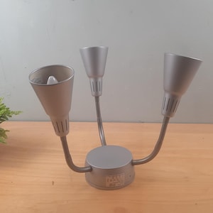 May include: A gray, three-light fixture with adjustable, cone-shaped shades. The fixture has a circular base and flexible arms. The light is on a wooden surface.