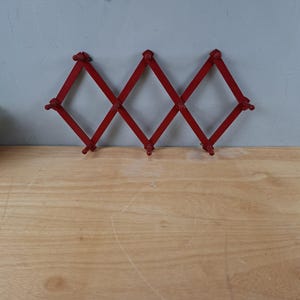 Könnte beinhalten: Ein roter, ausziehbarer Wandhaken aus Holz im Akkordeon-Stil mit einem Gitterdesign und mehreren Stiften zum Aufhängen von Gegenständen. Das Gestell besteht aus miteinander verbundenen Leisten, die ein Rautenmuster bilden.