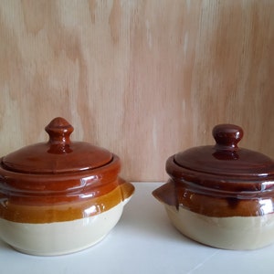 May include: Two brown and beige ceramic casserole dishes with lids. The dishes have a rounded shape and two small handles on the sides.