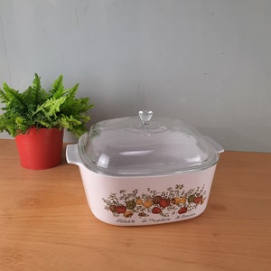 Puede incluir: Plato para hornear de cerámica blanca con tapa de vidrio. El plato tiene un diseño floral con acentos rojos, amarillos y verdes. El texto "L'Echalote La Marjolaine Le Romarin" está impreso en la parte inferior del plato.