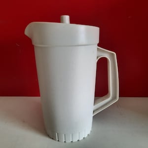 Può includere: Brocca di plastica bianca con coperchio e manico. La brocca ha un manico rettangolare e un beccuccio per versare. Il coperchio ha un piccolo pomello sulla parte superiore. La brocca è su una superficie bianca su sfondo rosso.