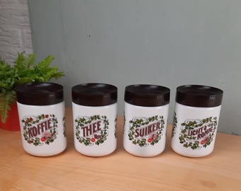 Op de afbeelding: Vier witte keramische potten met donkerbruine deksels. Elke pot heeft een decoratief etiket met tekst en bloemmotieven. De etiketten luiden "KOFFIE", "THEE", "SUIKER" en "LICHT & ROMIG".