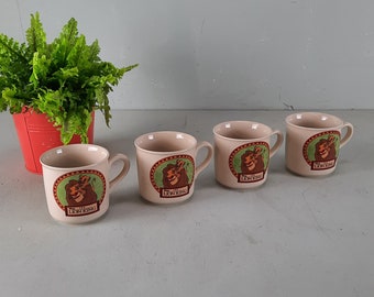Może przedstawiać: Zestaw czterech ceramicznych kubków z kreskówkowym wizerunkiem Simby i Timona z filmu Disneya Król Lew. Kubki są beżowe z zielonym, czerwonym i brązowym wzorem.