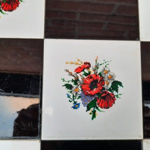 May include: A white tile with a black border features a floral design with red, white, and blue flowers. The flowers are arranged in a bouquet and are surrounded by green leaves.