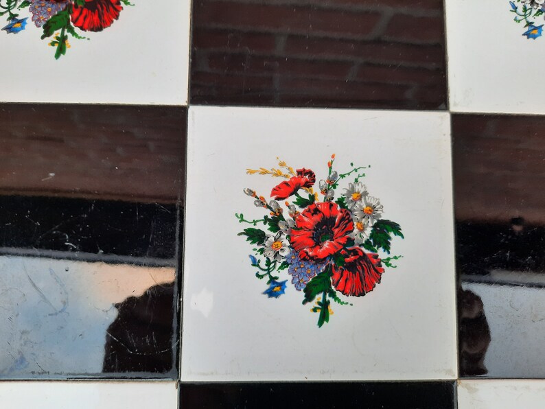May include: A white tile with a black border features a floral design with red, white, and blue flowers. The flowers are arranged in a bouquet and are surrounded by green leaves.