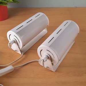 Peut inclure: Deux lampes cylindriques blanches avec un cordon et une prise blancs. Les lampes ont une base en métal blanc et un abat-jour en plastique blanc avec une série de fentes.