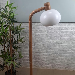 Może przedstawiać: Drewniana lampa podłogowa z białym, kulistym kloszem. Lampa ma zakrzywione, segmentowane ramię i okrągłą podstawę. Drewniane elementy są jasnobrązowe, a lampa stoi przed białą ceglaną ścianą.