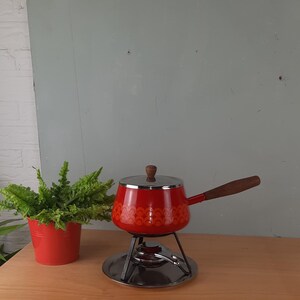 Op de afbeelding: Rode emaille fonduepot met een houten handvat en een patroon. De pot staat op een metalen standaard met een brander.