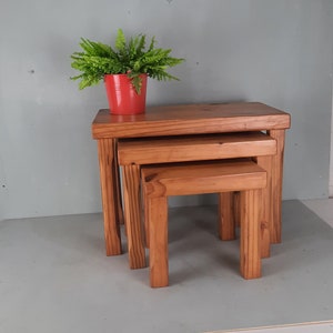 Puede incluir: Un conjunto de tres mesas nido de madera en un acabado marrón cálido. Las mesas están apiladas una encima de la otra, con la mesa más grande en la parte inferior y la mesa más pequeña en la parte superior. Una pequeña planta verde en maceta se coloca sobre la mesa superior.