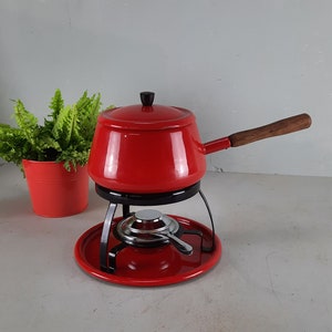Puede incluir: Una olla de fondue roja con un soporte negro y un asa de madera. La olla se asienta sobre una base roja con un quemador. Una pequeña olla roja con una planta verde está en el fondo.