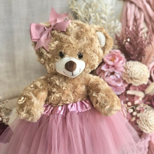 May include: A tan plush teddy bear with a textured, curly fur finish. The bear features a dark brown nose, black eyes, and a pink ribbon bow attached to its head. It is wearing a layered pink tulle skirt with a satin waistband around its middle.