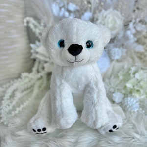 May include: White plush polar bear stuffed animal with blue eyes and a black nose. The bear is sitting on a white fluffy surface.