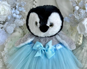 May include: A plush penguin toy wearing a light blue tulle tutu with a matching satin bow. The penguin has black and white fur and a black beak.