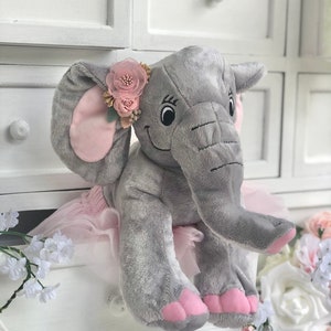 May include: A gray plush elephant wearing a pink tutu and a pink flower crown. The elephant has pink ears and pink feet.