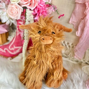 May include: A plush brown Highland cow toy with a fluffy coat. The toy is sitting on a white fluffy surface.