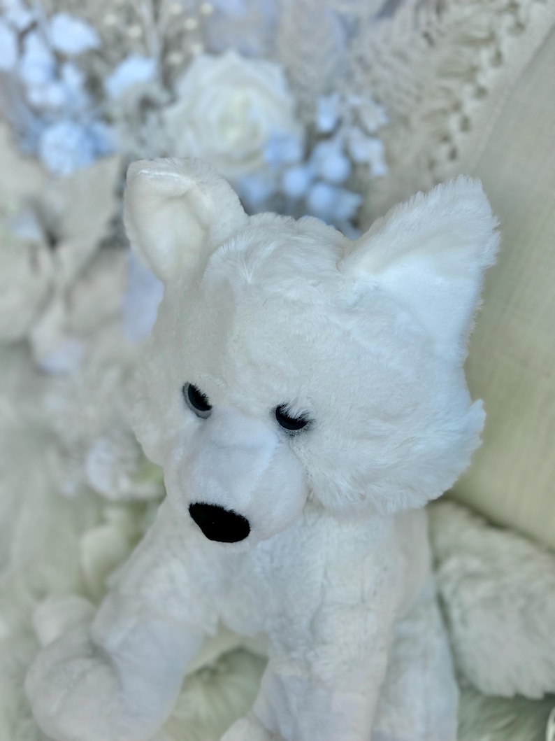 K&ouml;nnte beinhalten: Ein wei&szlig;es Pl&uuml;schtier in Form eines Eisb&auml;ren mit schwarzen Augen und einer schwarzen Nase. Das Kuscheltier hat gro&szlig;e Ohren und sitzt auf einer wei&szlig;en Oberfl&auml;che. Der Hintergrund zeigt wei&szlig;e und hellblaue Blumendekorationen.