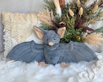 Könnte beinhalten: Ein Fledermaus-Plüschtier mit dunkelgrauen Flügeln und Körper, hellbraunen Innenohren und Füßen, einer kleinen beigen Nase und einem strukturierten grauen Kragen. Die Fledermaus hat große, dunkle Augen und ist mit ausgebreiteten Flügeln dargestellt.